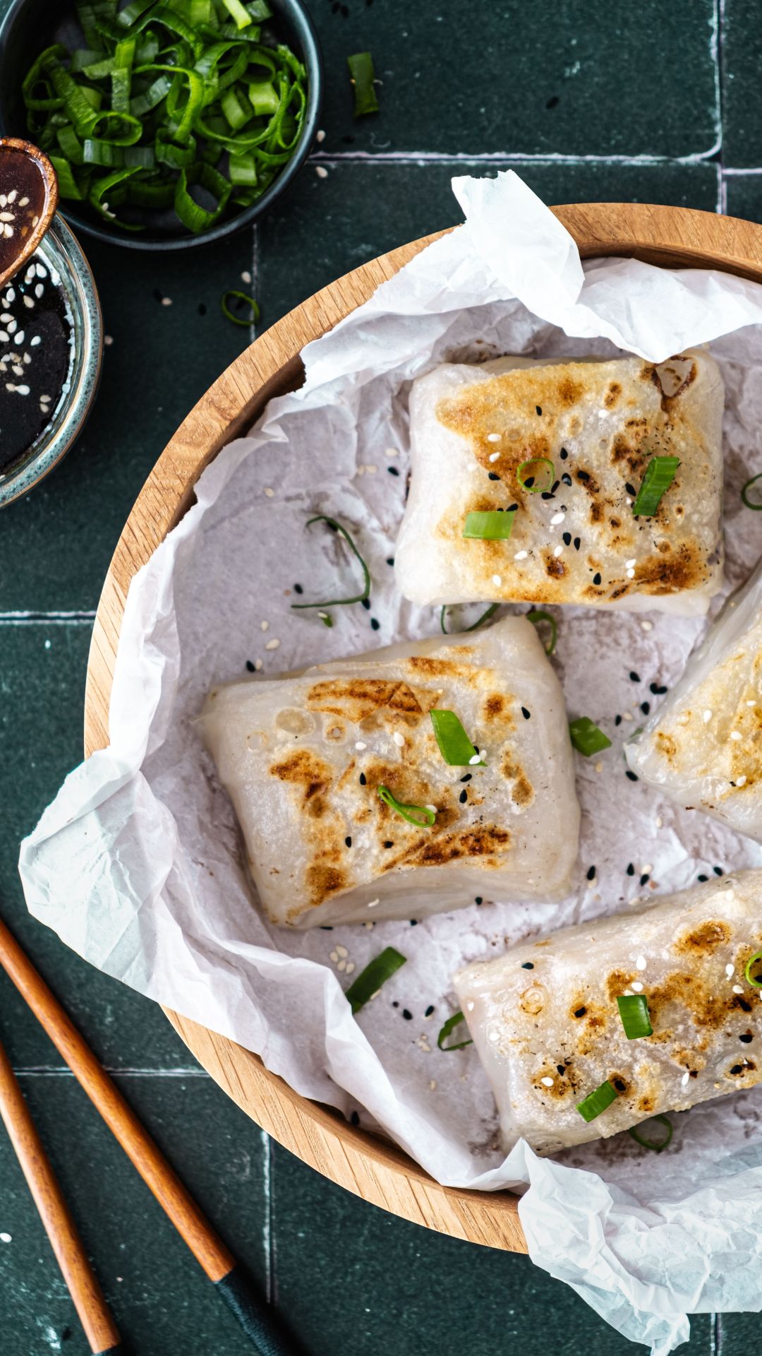 Knusprige Reispapier Dumplings in einer Holzschale. Ein Schälchen mit Honig-Sauce und einem Holzlöffel. Essstäbchen aus Holz. Petersilie-Blätter und ein Schälchen mit Frühlingszwiebel-Ringe.