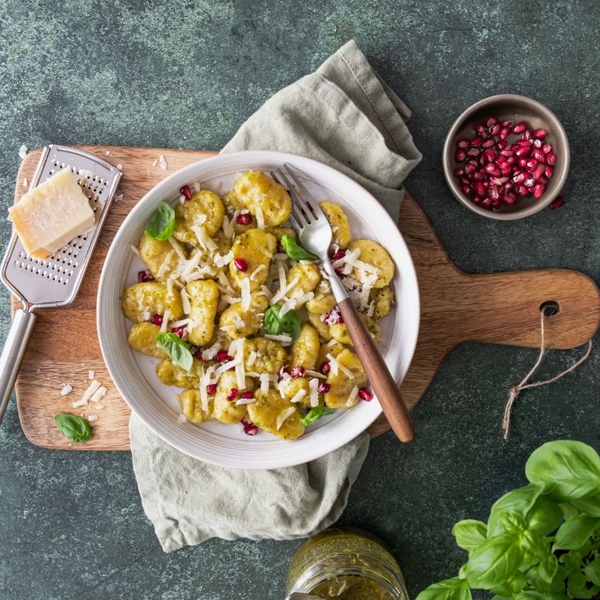 Sellerie-Gnocchi im Teller auf einem Holzbrett. Parmesankäse mit Reibe, Pistazienpesto, Basilikumtopf und Granatapfelkerne als Deko