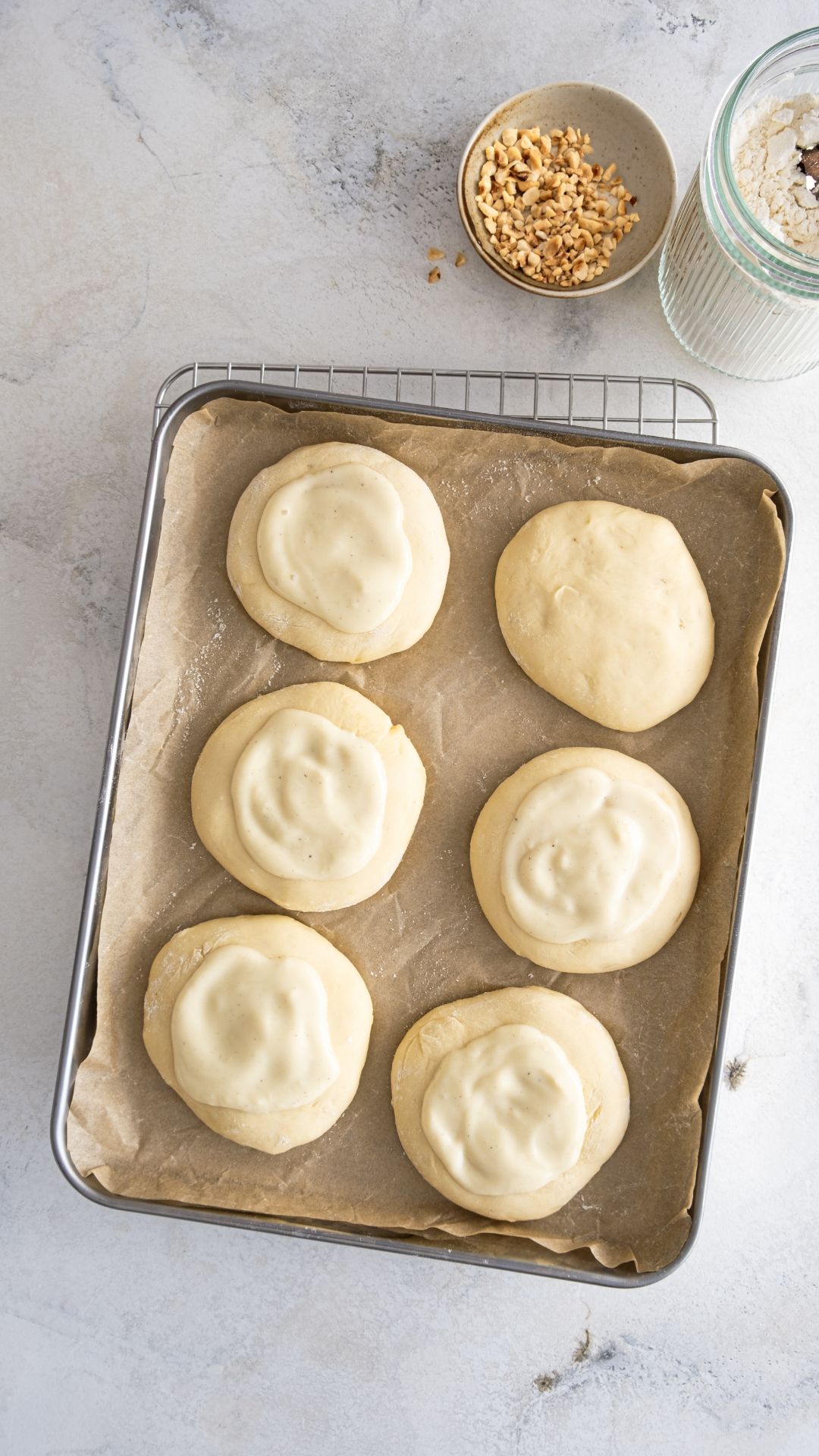 Sechs Hefeteiglinge auf einem Backblech, mit einer cremigen Schicht veganer Füllung bestrichen. Im Hintergrund eine Schale gehackten Nüssen und ein Glas Mehl.