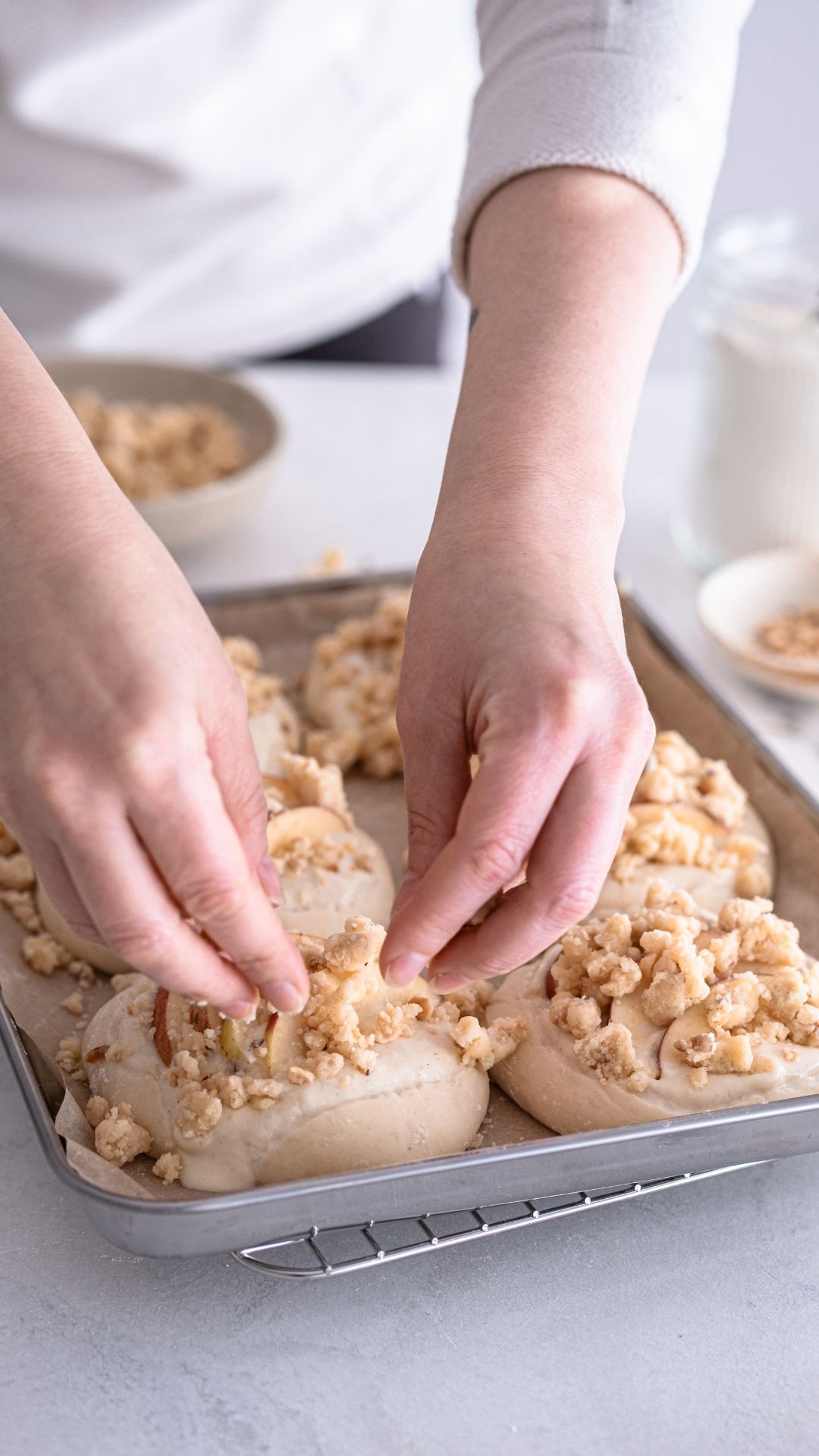 Sechs Hefeteiglinge auf einem Backblech, mit einer cremigen Schicht veganer Füllung bestrichen und mit Apfelscheiben belegt. Die Streusel werden darüber gestreut. Im Hintergrund eine Schale gehackten Nüssen und ein Glas Mehl.